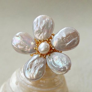 May include: A white pearl flower ring with a gold centre and a single pearl in the centre. The ring is made of freshwater pearls and is a delicate and elegant piece of jewellery.