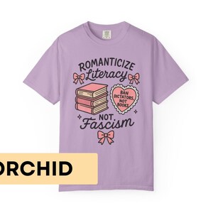 May include: Orchid-colored t-shirt with the text "Romanticize Literacy, Not Fascism." The design includes a stack of books, bows, and a heart-shaped graphic that reads "Ban Dictators, Not Books."