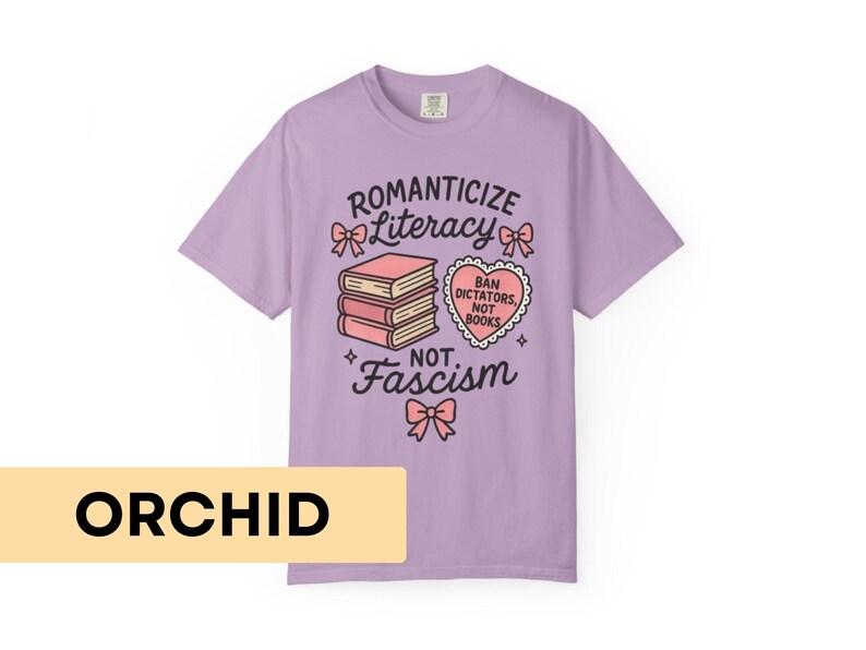 May include: Orchid-colored t-shirt with the text "Romanticize Literacy, Not Fascism." The design includes a stack of books, bows, and a heart-shaped graphic that reads "Ban Dictators, Not Books."
