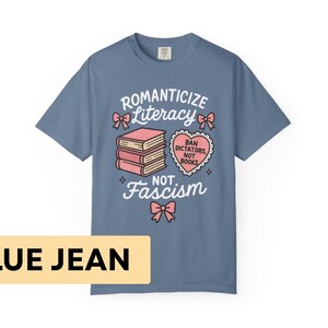 May include: Blue jean colored t-shirt with the words "Romanticize Literacy, Not Fascism" in white script. The design includes pink bows, a stack of books, and a heart-shaped graphic that reads "Ban Dictators, Not Books."