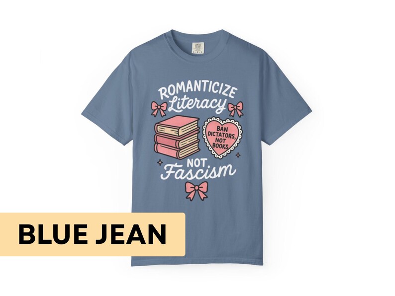 May include: Blue jean colored t-shirt with the words "Romanticize Literacy, Not Fascism" in white script. The design includes pink bows, a stack of books, and a heart-shaped graphic that reads "Ban Dictators, Not Books."