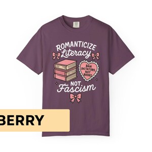 May include: Berry-colored t-shirt with a graphic design. The design includes the words "Romanticize Literacy, Not Fascism" with illustrations of books and a heart-shaped graphic. The shirt is made of a soft material.