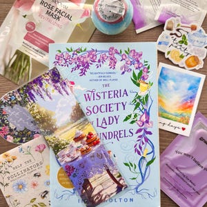 May include: A flat lay of various items including a book titled "The Wisteria Society Lady Indrels" by I.K. Holton, a bath bomb, a face mask, a bookmark, a tea bag, and a sticker.
