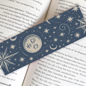 May include: A celestial-themed bookmark with a navy blue background. It features a crescent moon with a face, stars, and a compass rose design. The bookmark is rectangular and made of a clear, glossy material.