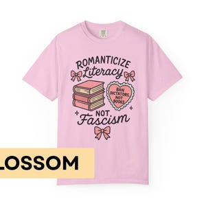 May include: A light pink t-shirt with the words "Romanticize Literacy Not Fascism" and a graphic of books and a heart. The shirt also has the word "Blossom" in a yellow rectangle at the bottom.