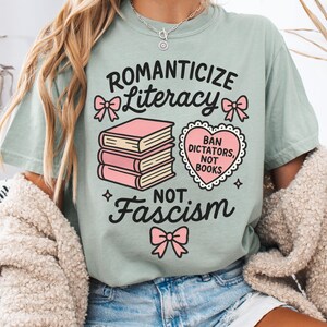 May include: A light green t-shirt with the words "Romanticize Literacy, Not Fascism" in black script. The shirt features pink bow accents and a stack of books. A heart-shaped graphic reads "Ban Dictators, Not Books."