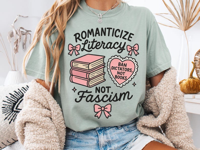 May include: A light green t-shirt with the words "Romanticize Literacy, Not Fascism" in black script. The shirt features pink bow accents and a stack of books. A heart-shaped graphic reads "Ban Dictators, Not Books."