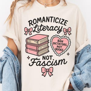 May include: Cream-colored t-shirt with the words "Romanticize Literacy, Not Fascism" in black script. The design includes pink bows, a stack of pink books, and a heart-shaped graphic that reads "Ban Dictators, Not Books."