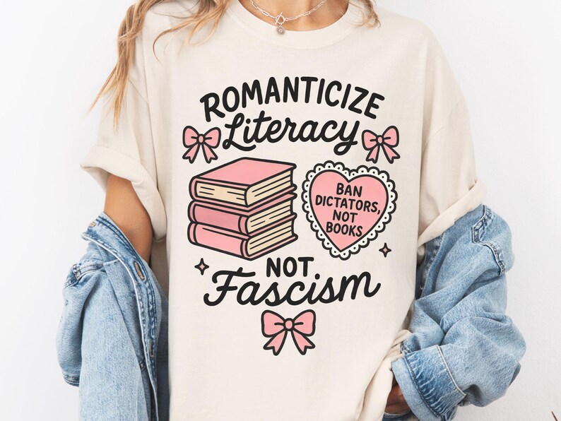 May include: Cream-colored t-shirt with the words "Romanticize Literacy, Not Fascism" in black script. The design includes pink bows, a stack of pink books, and a heart-shaped graphic that reads "Ban Dictators, Not Books."