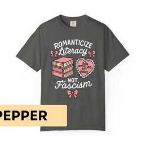 May include: Dark gray t-shirt with the text "Romanticize Literacy, Not Fascism." The design includes pink bows, a stack of books, and a heart-shaped graphic that reads "Ban Dictators, Not Books."