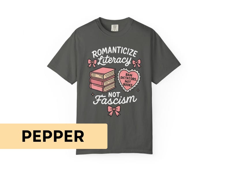 May include: Dark gray t-shirt with the text "Romanticize Literacy, Not Fascism." The design includes pink bows, a stack of books, and a heart-shaped graphic that reads "Ban Dictators, Not Books."