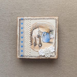 May include: A small wooden square with a brown stitched border. The centre features a vintage illustration of three bunnies and a woman in a blue dress. The background is a light brown with a white lace trim. A white flower with a pearl centre is attached to the bottom right corner.