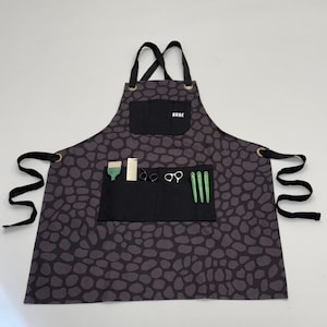 May include: Black and grey patterned apron with a black pocket and the word "RUDE" printed on it. The apron has adjustable straps and a variety of tools including scissors, combs, and clips.