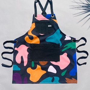 Op de afbeelding: Een zwarte schort met een kleurrijk abstract patroon. De schort heeft een grote zwarte zak met het woord "RUDE" in wit erop gedrukt. De schort heeft verstelbare banden en touwtjes.