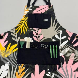 May include: Black apron with a tropical leaf print in shades of pink, yellow, white, and grey. The apron has a large black pocket with the word "RUDE" printed in white. The pocket contains a green comb, a pair of silver scissors, and three green hair clips.