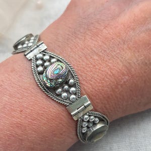 Può includere: Un bracciale articolato color argento con un inserto centrale in conchiglia di abalone iridescente di forma ovale. La lavorazione del metallo include dettagli decorativi a perline e bordi a corda intrecciata attorno alla pietra centrale.