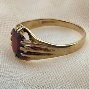 May include: A gold ring featuring a rectangular, deep red gemstone set in a prong setting. The band has a smooth, polished finish and a split-shank design that connects to the central stone setting, showcasing a classic, metallic construction.