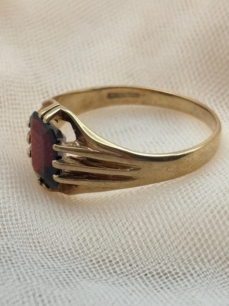 May include: A gold ring featuring a rectangular, deep red gemstone set in a prong setting. The band has a smooth, polished finish and a split-shank design that connects to the central stone setting, showcasing a classic, metallic construction.