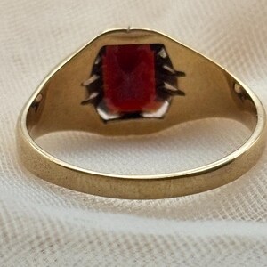 May include: A gold ring featuring a rectangular, deep red gemstone set in a secure metal mounting. The band is smooth and circular, showcasing a polished gold finish that contrasts with the vibrant, dark red stone held by visible prongs.