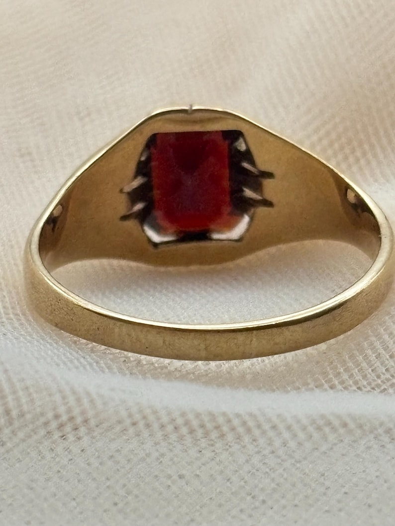 May include: A gold ring featuring a rectangular, deep red gemstone set in a secure metal mounting. The band is smooth and circular, showcasing a polished gold finish that contrasts with the vibrant, dark red stone held by visible prongs.