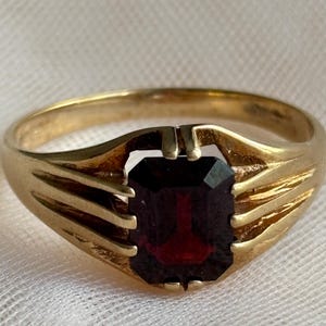 May include: A gold ring featuring a rectangular, emerald-cut dark red gemstone set in a prong setting. The band is crafted from polished yellow gold and displays a split-shank design that tapers toward the center stone, creating a structured look.