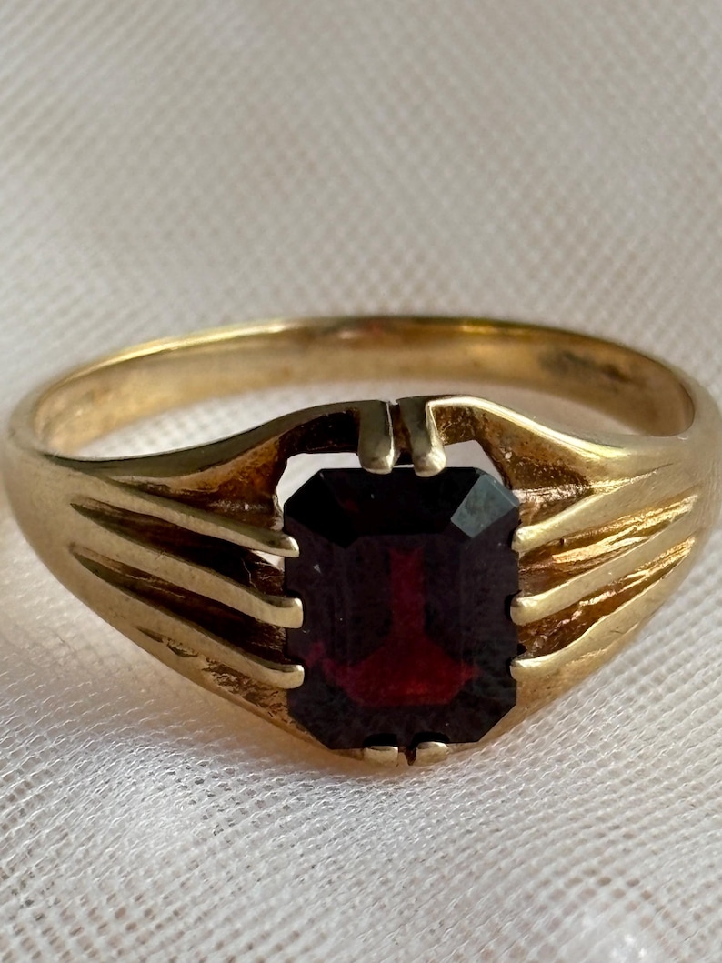 May include: A gold ring featuring a rectangular, emerald-cut dark red gemstone set in a prong setting. The band is crafted from polished yellow gold and displays a split-shank design that tapers toward the center stone, creating a structured look.
