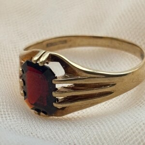 May include: A gold ring featuring a rectangular, deep red gemstone set in a prong setting. The band is crafted from polished gold and displays a multi-pronged design that secures the stone, with a smooth, tapered shank extending around the finger.