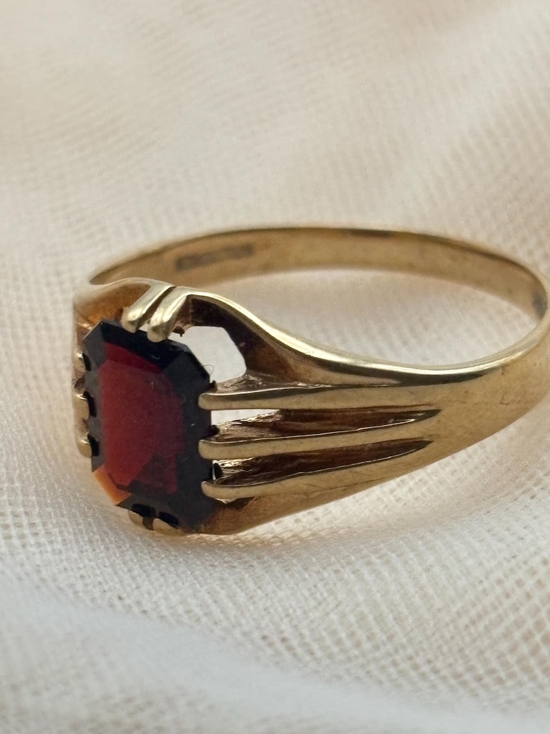 May include: A gold ring featuring a rectangular, deep red gemstone set in a prong setting. The band is crafted from polished gold and displays a multi-pronged design that secures the stone, with a smooth, tapered shank extending around the finger.