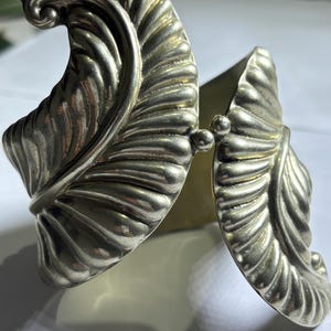 May include: Silver cuff bracelet with a leaf design. The bracelet features detailed, textured leaves that curve around the wrist. The leaves are connected by a hinge with two small, round beads.
