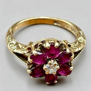 May include: A gold ring featuring a floral cluster design with six oval-cut deep red rubies surrounding a single central round-cut clear diamond. The gold band displays intricate engraved leaf patterns along the sides of the setting.