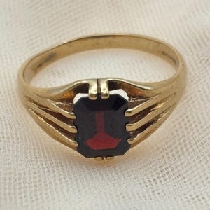 May include: A gold ring featuring a rectangular, dark red gemstone set in the center. The band is constructed with three distinct, parallel gold prongs on each side that extend upward to secure the stone, creating an open, layered design.