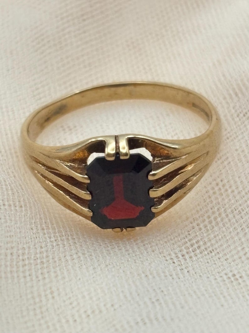 May include: A gold ring featuring a rectangular, dark red gemstone set in the center. The band is constructed with three distinct, parallel gold prongs on each side that extend upward to secure the stone, creating an open, layered design.