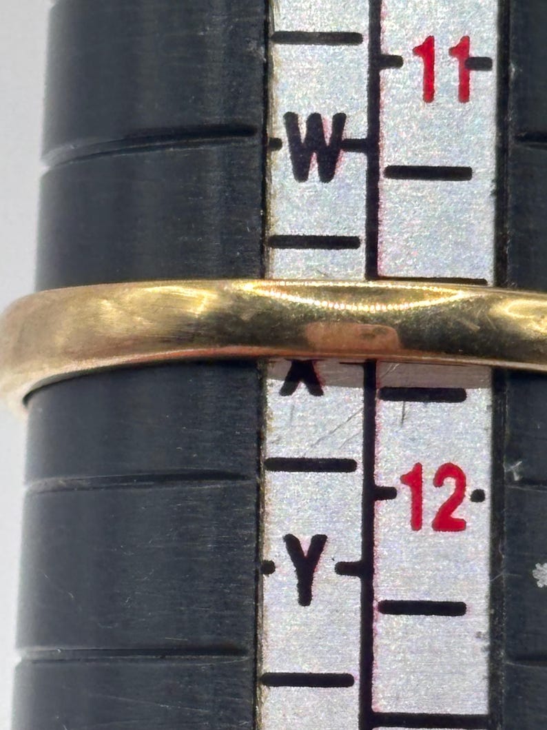 May include: A gold ring band positioned on a black ring sizing mandrel. The ring rests between the size markings W and Y on the vertical scale, indicating a size that aligns with the 11 and 12 markers printed in red on the white measuring tool.