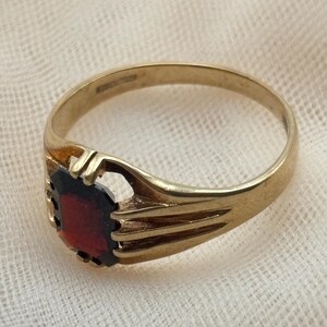 May include: A gold ring featuring a rectangular, deep red gemstone set in a pronged mounting. The band is smooth and tapers toward the setting, which includes open, decorative metalwork on the sides to secure the central stone in place.