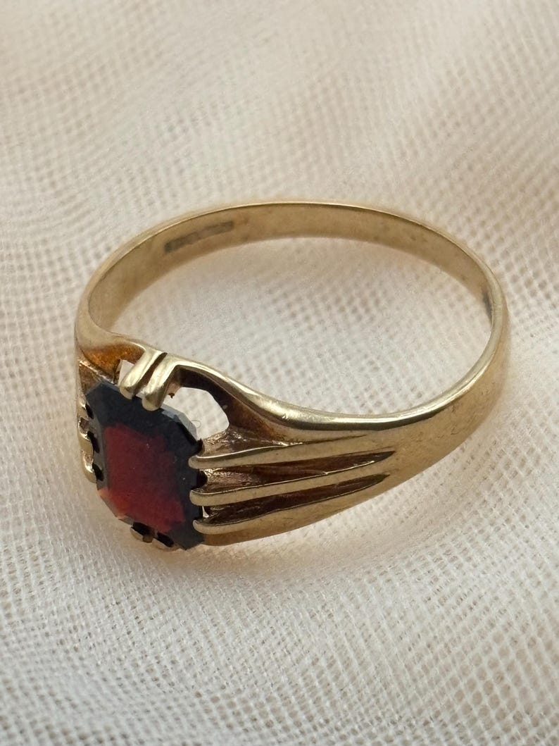 May include: A gold ring featuring a rectangular, deep red gemstone set in a pronged mounting. The band is smooth and tapers toward the setting, which includes open, decorative metalwork on the sides to secure the central stone in place.