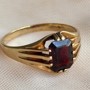 May include: A gold ring featuring a rectangular, emerald-cut dark red gemstone set in a prong setting. The band is crafted from polished gold and displays a split-shank design where the metal divides into three distinct prongs on each side.