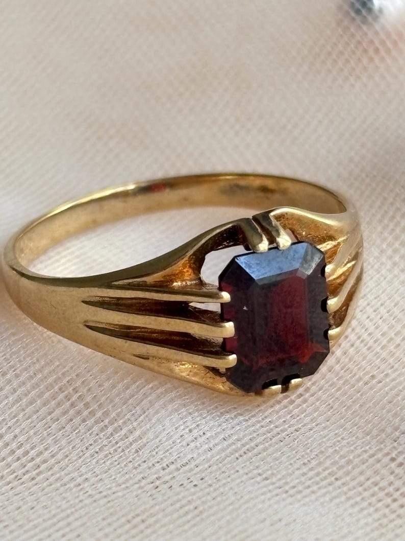May include: A gold ring featuring a rectangular, emerald-cut dark red gemstone set in a prong setting. The band is crafted from polished gold and displays a split-shank design where the metal divides into three distinct prongs on each side.