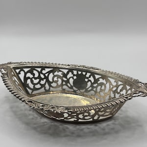 May include: A silver-coloured, oblong decorative basket with intricate openwork filigree patterns. The basket has a beaded edge and a smooth, reflective interior. The design is symmetrical, with a small emblem visible on the front.