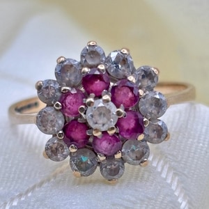 Puede incluir: Un anillo de oro con un diseño de racimo floral. El centro es una piedra clara rodeada por seis rubíes redondos de color rosa intenso. Un halo exterior de doce piedras claras rodea los rubíes, todo montado en una banda de oro pulido.