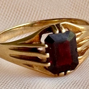 May include: A gold ring featuring a rectangular, deep red gemstone set in the center. The band is crafted from polished gold and displays a series of three parallel, raised decorative lines on each side that extend toward the central stone setting.