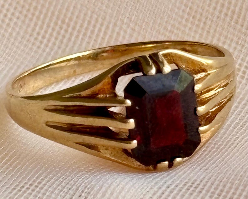 May include: A gold ring featuring a rectangular, deep red gemstone set in the center. The band is crafted from polished gold and displays a series of three parallel, raised decorative lines on each side that extend toward the central stone setting.