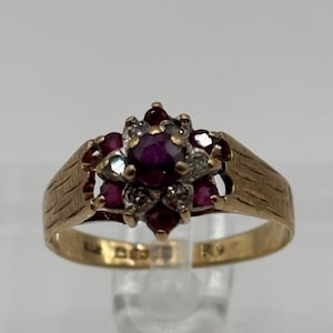 May include: A gold ring with a central ruby surrounded by smaller rubies and clear gemstones. The ring's band has a textured pattern. The ring is a classic piece of jewellery.