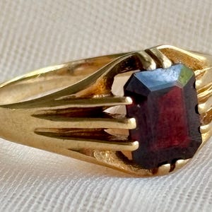 May include: A gold ring featuring a rectangular, deep red gemstone set in a prong setting. The band is crafted with a series of parallel, grooved lines that extend from the sides of the central stone, creating a structured, multi-ribbed design.