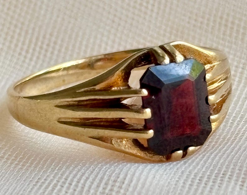 May include: A gold ring featuring a rectangular, deep red gemstone set in a prong setting. The band is crafted with a series of parallel, grooved lines that extend from the sides of the central stone, creating a structured, multi-ribbed design.