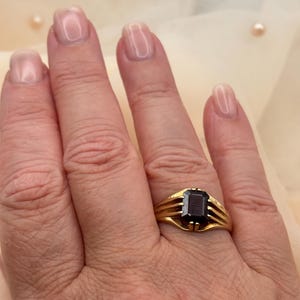May include: A gold ring featuring a rectangular, dark-colored faceted gemstone set in a prong setting. The band of the ring is composed of three thin, parallel gold wires that converge at the base of the stone, creating a structured design.