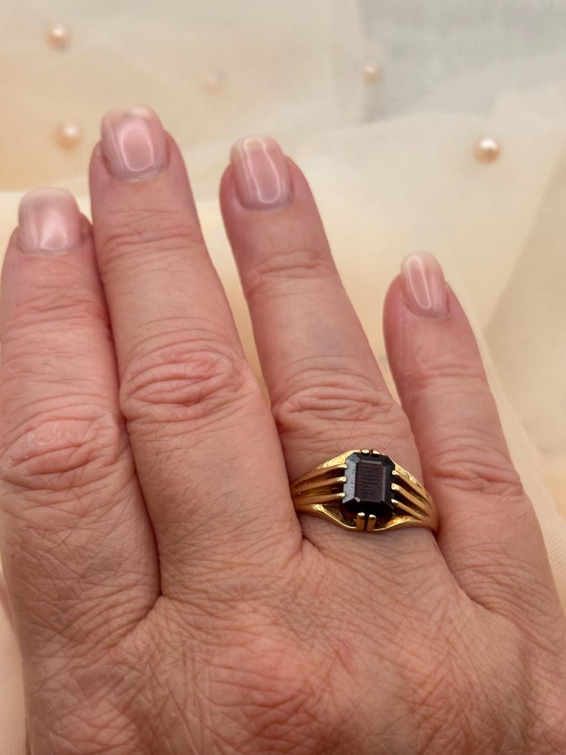 May include: A gold ring featuring a rectangular, dark-colored faceted gemstone set in a prong setting. The band of the ring is composed of three thin, parallel gold wires that converge at the base of the stone, creating a structured design.