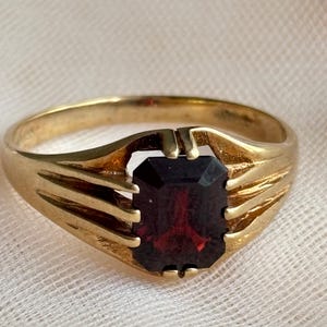 May include: A gold ring featuring a rectangular, deep red gemstone set in a prong setting. The band is crafted with a ribbed design on both sides, leading up to the central stone. The metal has a polished finish, highlighting the contrast between the gold and the dark red stone.