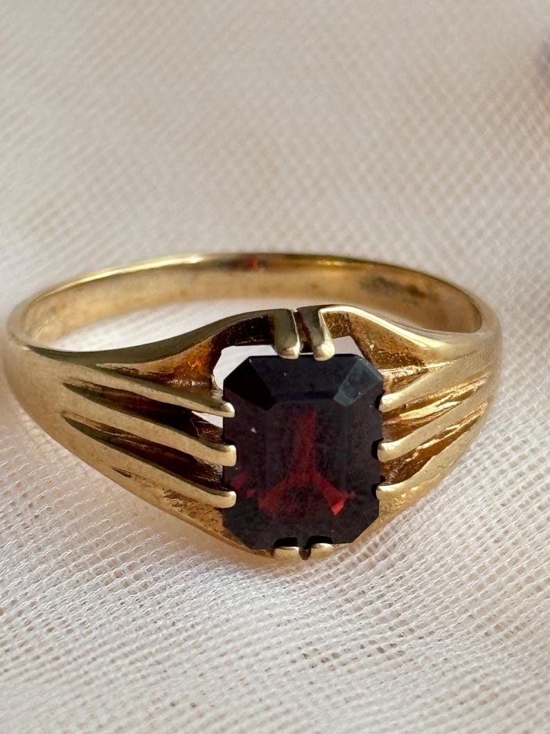 May include: A gold ring featuring a rectangular, deep red gemstone set in a prong setting. The band is crafted with a ribbed design on both sides, leading up to the central stone. The metal has a polished finish, highlighting the contrast between the gold and the dark red stone.