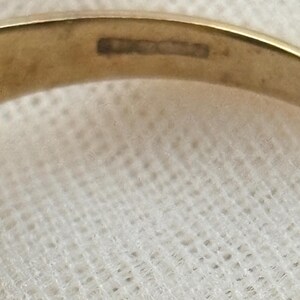 May include: A gold-colored metal ring featuring a smooth, polished band. The interior surface of the band displays a series of small, rectangular hallmarks or stamps, which are partially visible and slightly blurred, indicating metal purity or origin.