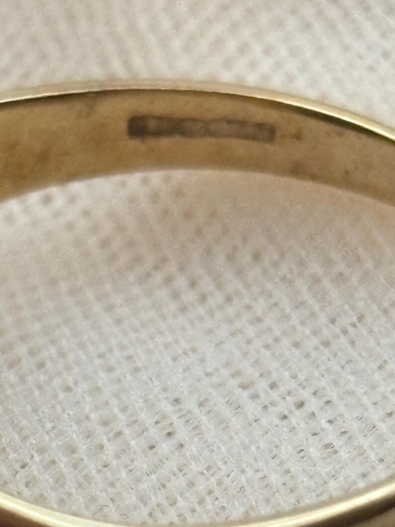 May include: A gold-colored metal ring featuring a smooth, polished band. The interior surface of the band displays a series of small, rectangular hallmarks or stamps, which are partially visible and slightly blurred, indicating metal purity or origin.